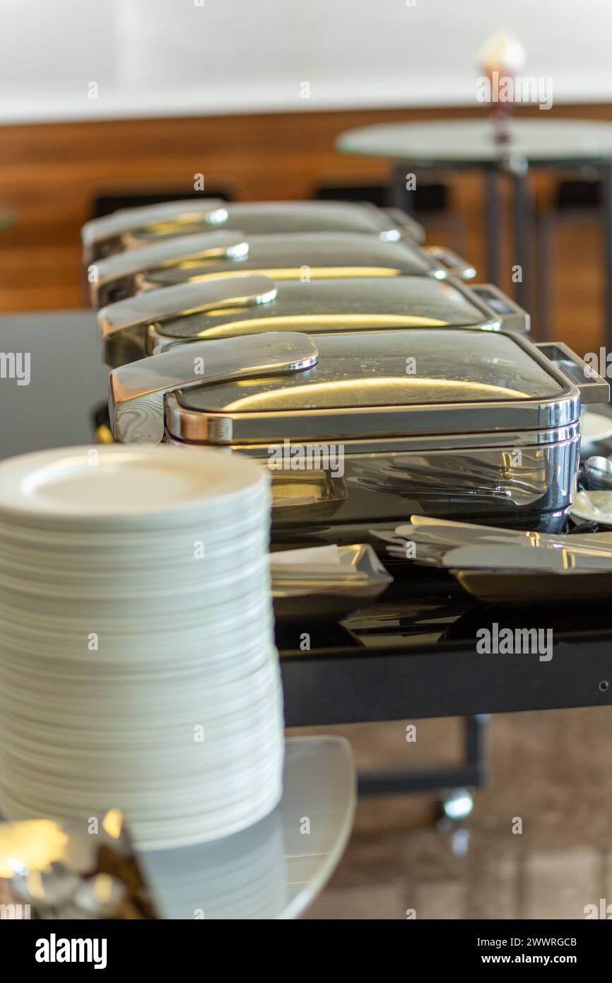 Pila di piatti accanto a piatti a base di chafing chiusi nella sala banchetti per feste. Marmites pronti per il servizio in acciaio inox a buffet. Foto Stock