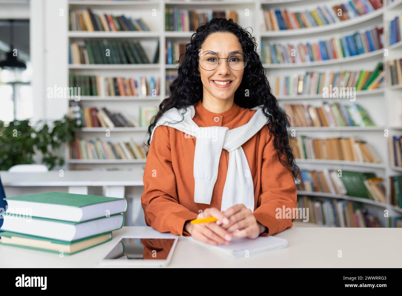 Tutor online contemporaneo seduto a tavola in biblioteca circondato da scaffali pieni di libri. Bella donna che indossa una giacca arancione con felpa con cappuccio bianca che tiene la penna gialla sul posto di lavoro personale. Foto Stock