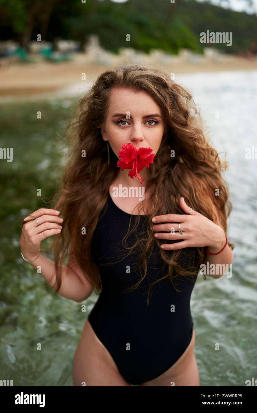 Una donna attraente tiene in bocca il fiore di ibisco rosso, si erge in costume da bagno nero in riva al mare. Bellezza femminile, vacanza tropicale, stile di vita da spiaggia, sensualità Foto Stock