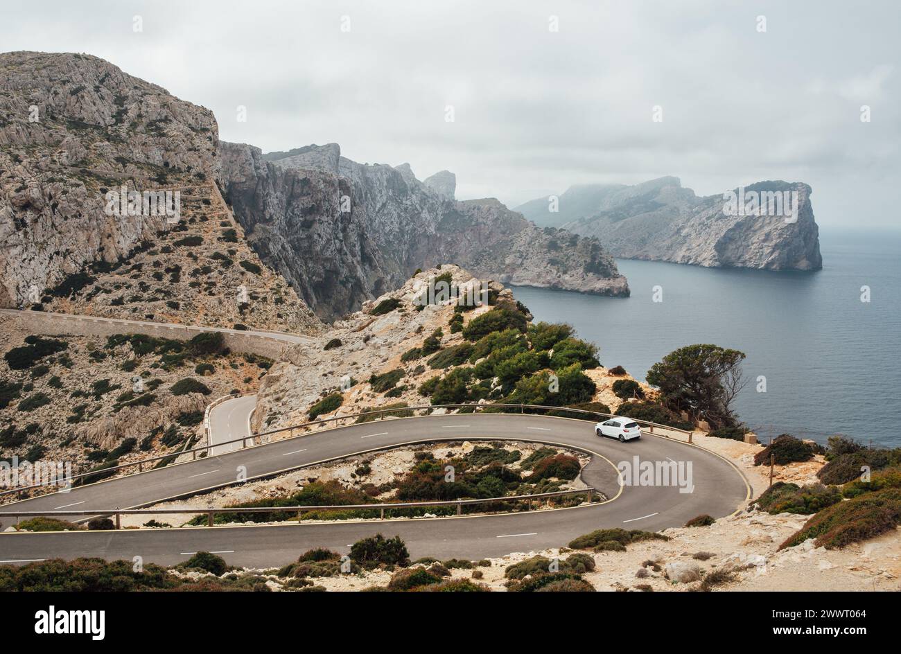 Piccola auto bianca solitaria che guida su una strada di montagna asfaltata curva a serpentina vicino al faro di Cap de Formentor con un bellissimo paesaggio marino con costa rocciosa Foto Stock