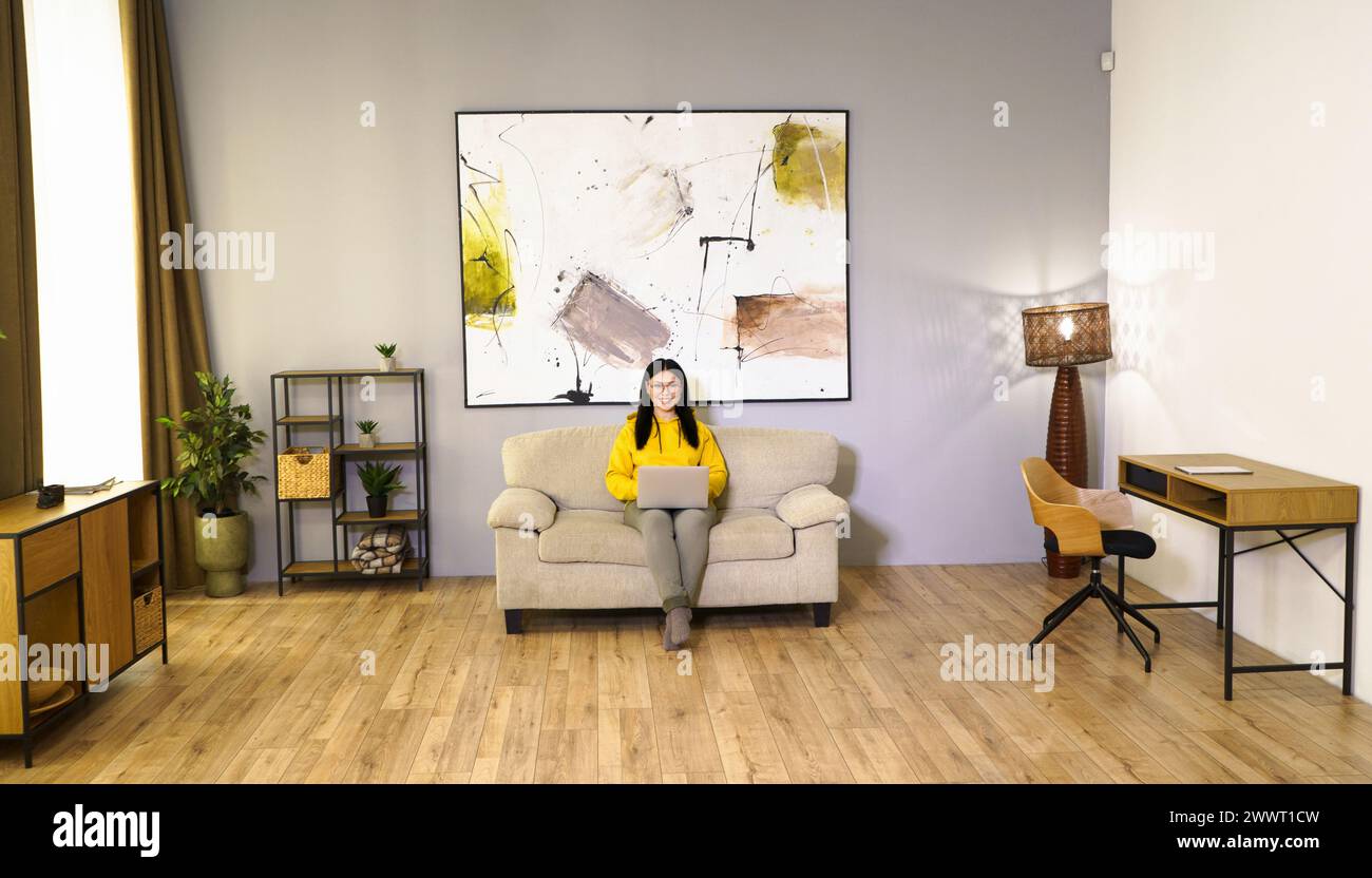 Una donna è seduta su un divano in un soggiorno con un portatile davanti a sé. La camera è decorata con un grande dipinto sulla parete e un pla in vaso Foto Stock