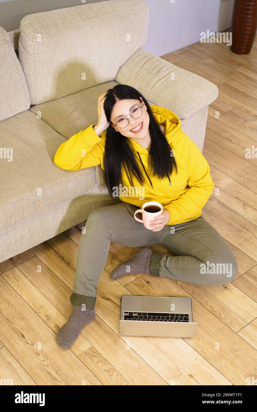Una donna con cappuccio giallo è seduta sul pavimento con un notebook e una tazza di caffè. Sta sorridendo e si sta godendo il suo tempo Foto Stock