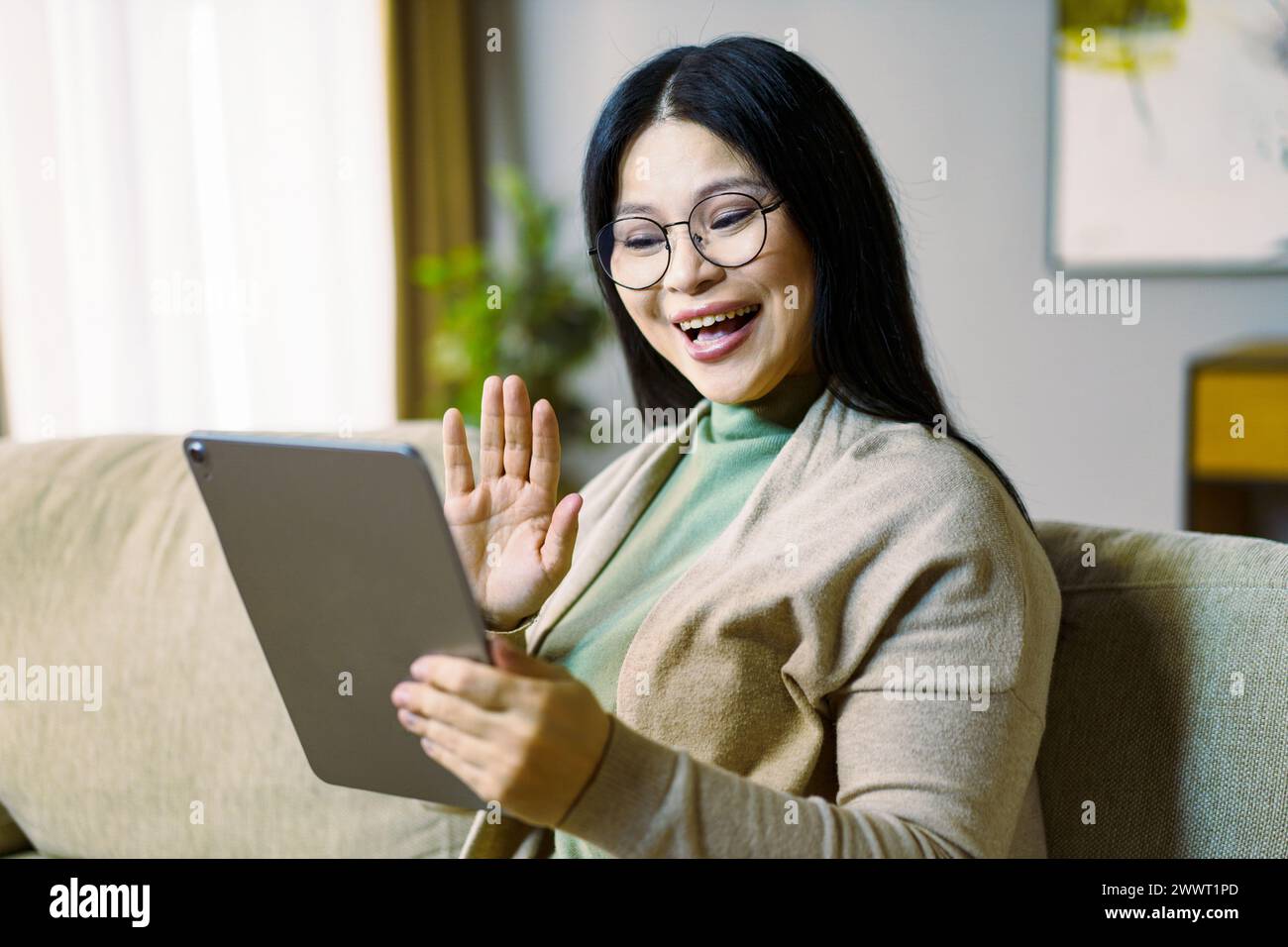 Una donna è seduta su un divano e sorride mentre tiene in mano un tablet. Sta salutando la telecamera, magari salutando qualcuno o esprimendo felicità. Conc Foto Stock