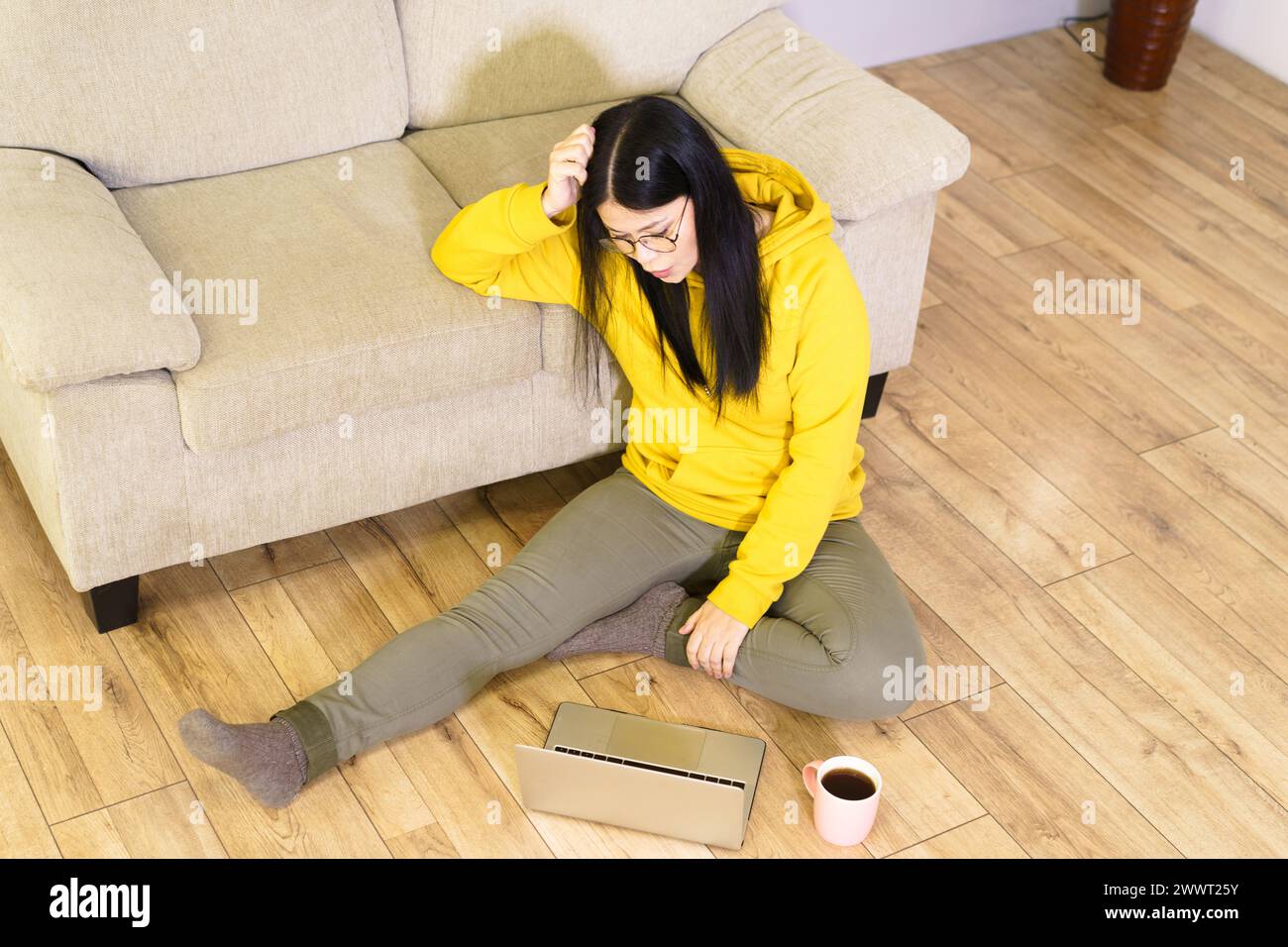 Una donna è seduta sul pavimento con un portatile di fronte a lei. Indossa una felpa gialla con cappuccio e indossa gli occhiali. La scena è informale e rilassata Foto Stock