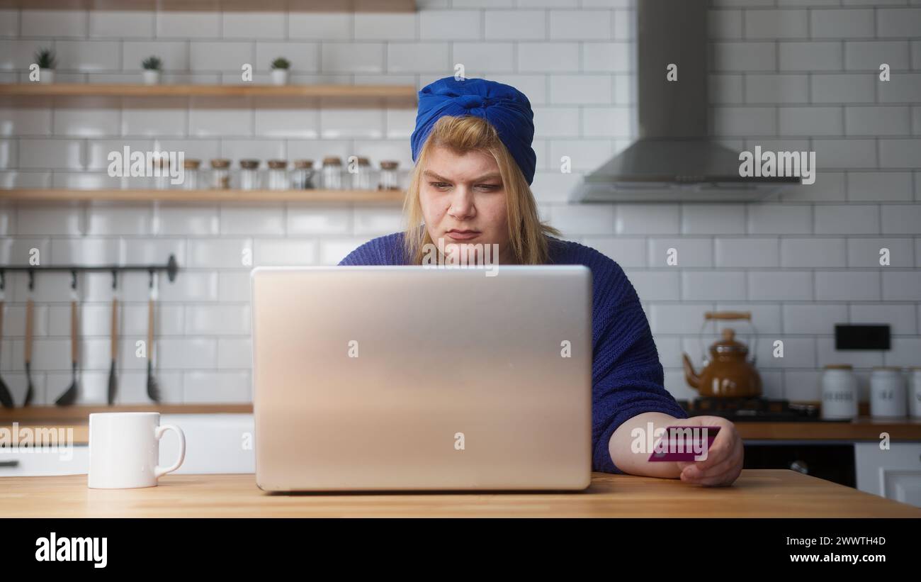 Una donna in sovrappeso immette il numero di carta di credito sul laptop per fare acquisti online, rende sicuro e facile il pagamento elettronico a distanza mentre è seduto in cucina a casa Foto Stock