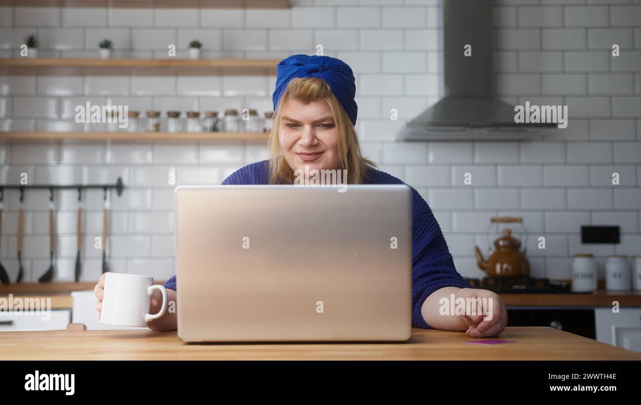 Una donna sorridente in sovrappeso inserisce il numero di carta di credito sul laptop per fare acquisti online, rende sicuro e facile il pagamento elettronico a distanza mentre si è seduti in cucina Foto Stock