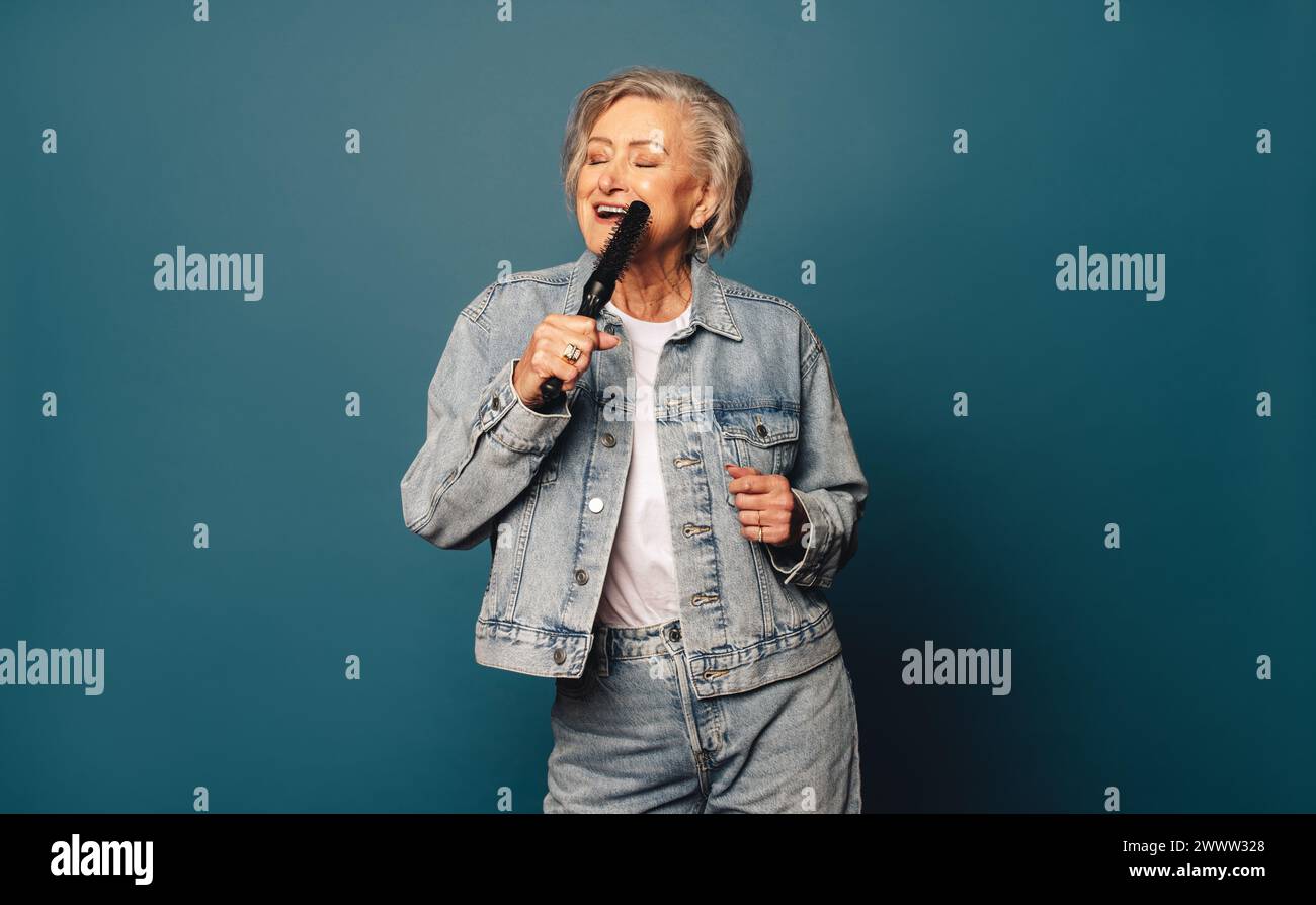Allegra donna anziana con capelli grigi balla e canta in uno studio. È vestita con un abbigliamento casual in denim e ha una spazzola per capelli. La sua giocosa e la sua cura Foto Stock