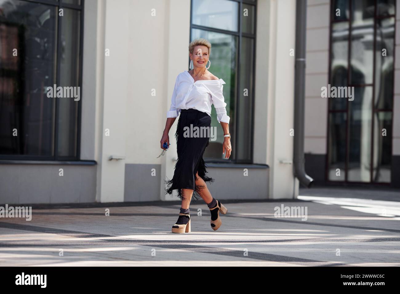 Fiducia e raffinatezza donna matura cammina per le strade della città in elegante ensemble camicia bianca chic e gonna con frange nere con sandalo con tacco alto Foto Stock