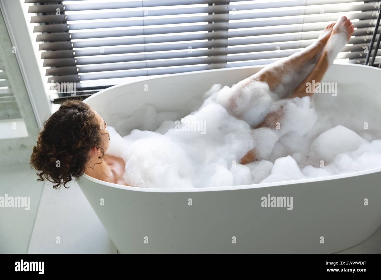 La donna si rilassa in un bagno di bolle a casa, con le gambe giocosamente sollevate Foto Stock