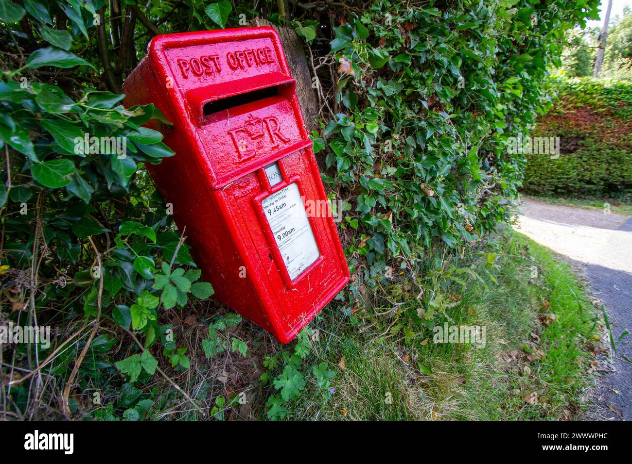 Sul lato di una strada di campagna, una cassetta delle lettere delle poste britanniche di colore rosso brillante si inclina in un angolo eccentrico Foto Stock