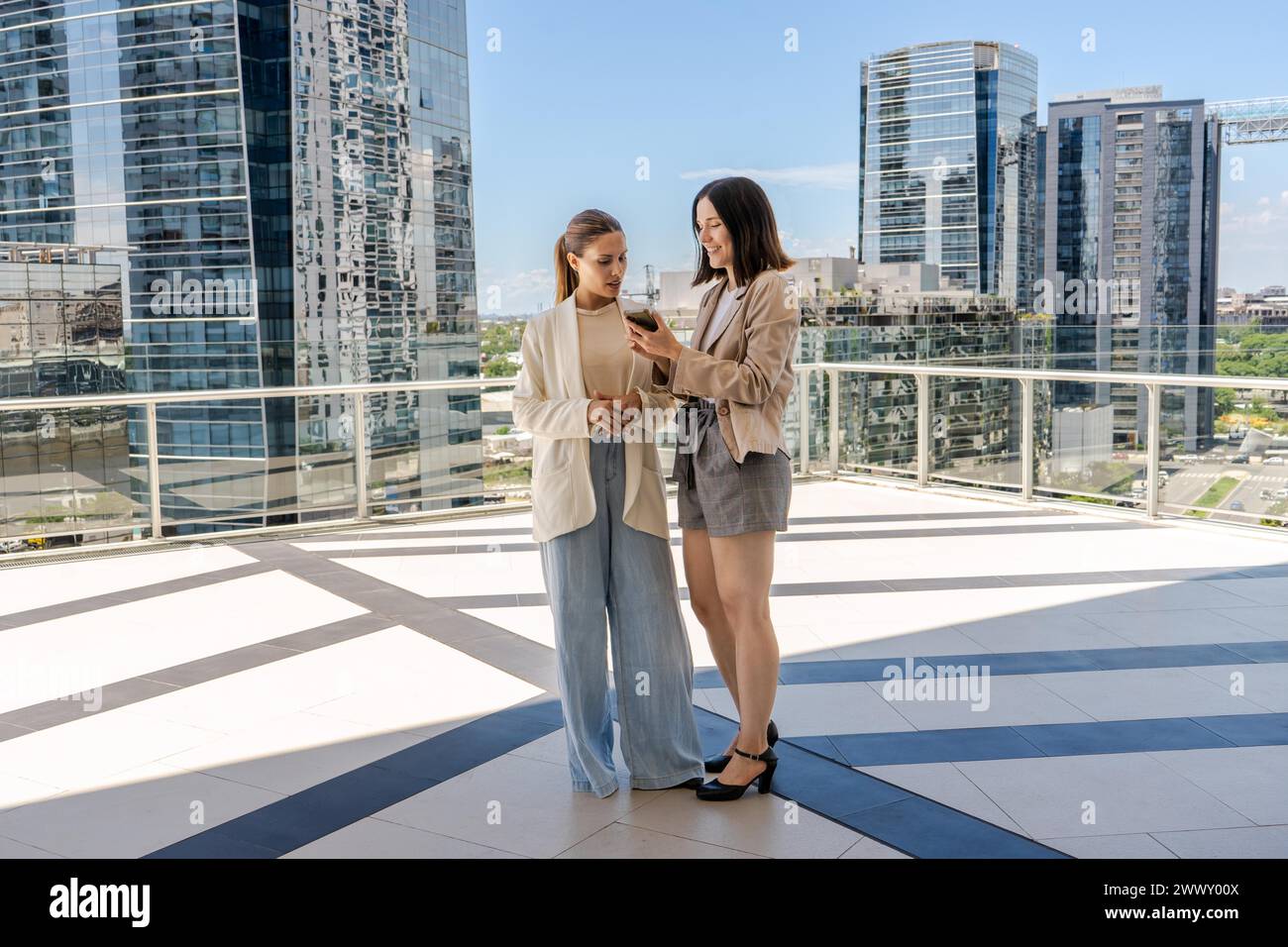 Due donne d'affari in piedi su una terrazza, una di loro tiene un cellulare e mostra un'e-mail al suo collega. La scena è ambientata in una città dall'alto Foto Stock