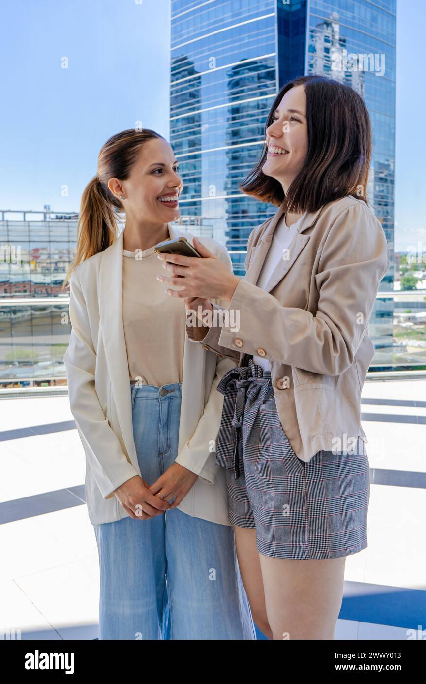 Due donne sono in piedi fuori, una di loro ha un cellulare. Stanno sorridendo e sembrano godersi la reciproca compagnia Foto Stock