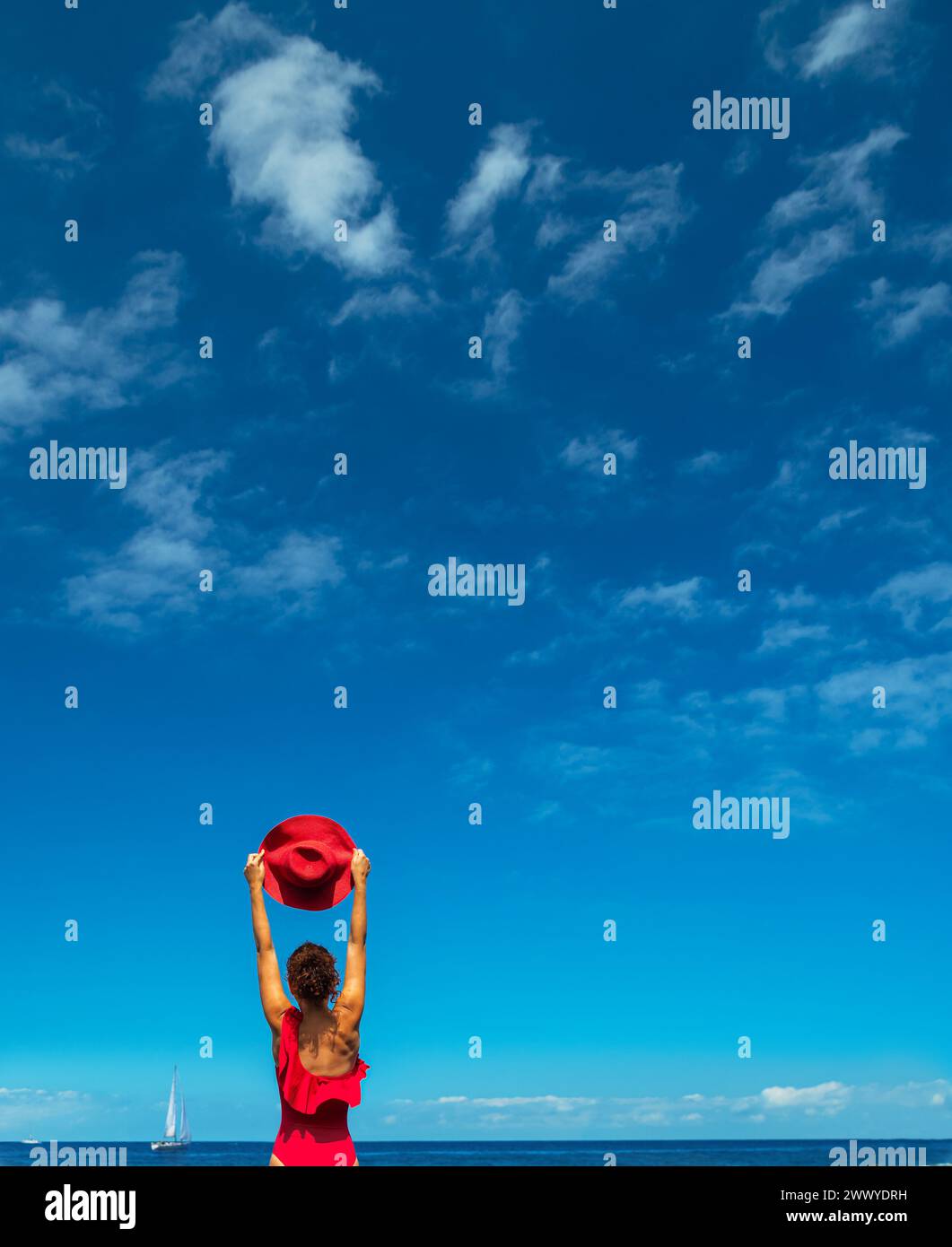 Una donna in costume da bagno rosso tiene il cappello rosso tra le mani. Calme acque oceaniche e cielo azzurro puro sullo sfondo. Foto Stock