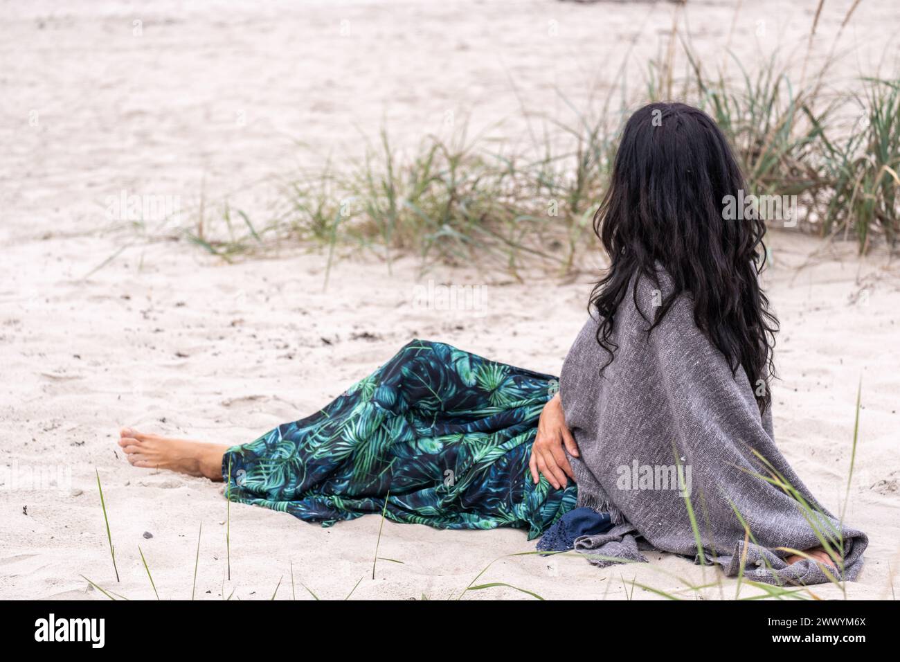 Donna contemplativa seduta sulla spiaggia, il suo sguardo perso nell'orizzonte, incarna un momento di riflessione pacifica ed equilibrio emotivo in mezza vita, come Foto Stock