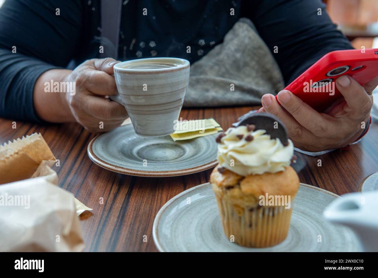 Primo piano di una persona irriconoscibile che beve una tazza di caffè con dolcificante e guarda il suo cellulare accanto a un dessert Foto Stock