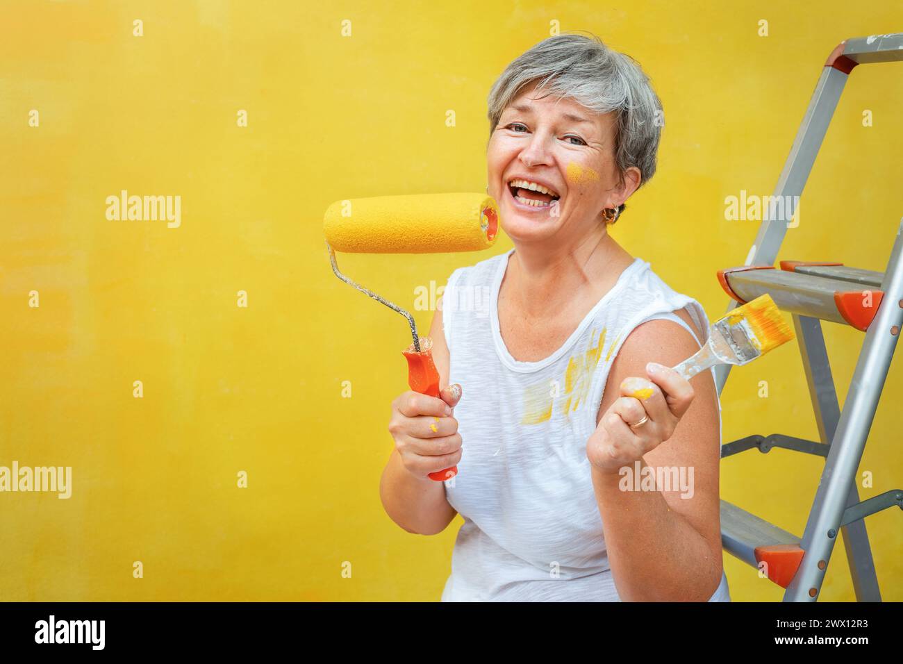 Senior felice seduto su una scala a cavalletto con un pennello e un rotolo di vernice vicino a una parete gialla Foto Stock
