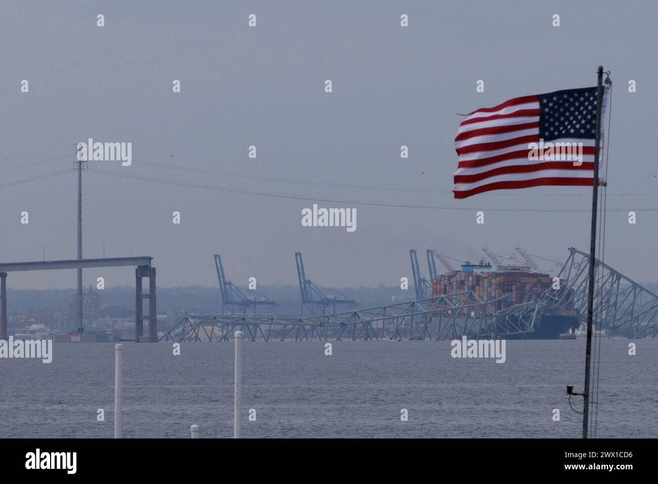 Una bandiera americana è visibile insieme ai resti del Francis Scott Key Bridge fuori Baltimora, Maryland, dopo che una nave portacontainer ha perso energia elettrica e ha colpito il ponte nelle prime ore del mattino martedì 26 marzo 2024 a Baltimora, MD, USA. Sei persone sono scomparse e presumibilmente morte dopo che una nave container ha colpito il monumento Francis Scott Key Bridge nella città americana di Baltimora. La Guardia Costiera ha detto di aver sospeso la ricerca e iniziato uno sforzo di recupero. Diversi veicoli stavano attraversando il ponte, che è lungo più di 1,6 km, quando è crollato dopo che la nave ha colpito un supporto. Dicono gli ufficiali Foto Stock