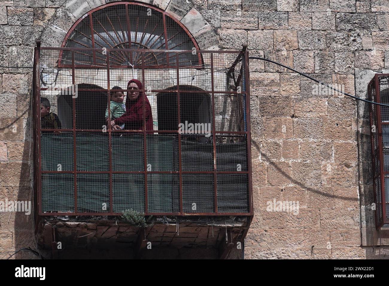 Una donna palestinese guarda con i suoi figli dal balcone della loro casa in via al-Shuhada, che è in gran parte chiusa ai palestinesi, durante l'annuale parata Purim mentre le forze di sicurezza israeliane si assicuravano le celebrazioni del 24 marzo 2024 a Hebron, Israele. Foto Stock