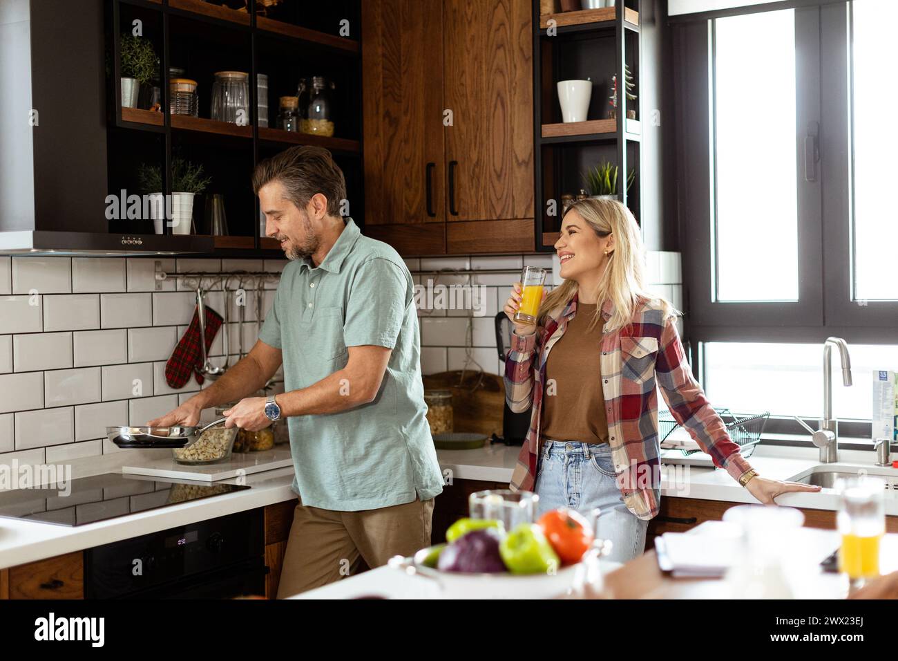 Un uomo e una donna che condividono una risata mentre cucinano in una cucina accogliente e ben organizzata Foto Stock