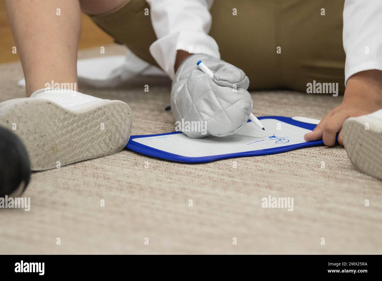 Primo piano di un bambino seduto in classe a giocare. Il ragazzo sta disegnando su una lavagna bianca con una sharpie mentre indossa un guanto da forno Foto Stock