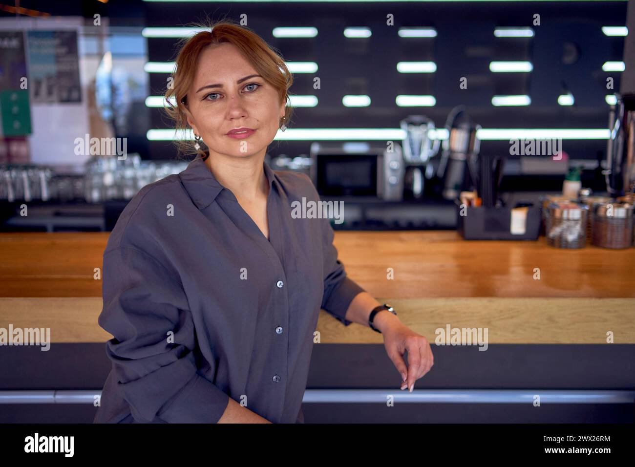 un'attraente donna di mezza età in un bar dal design neutro, che indossa pantaloni a gamba larga e una blusa in seta Foto Stock