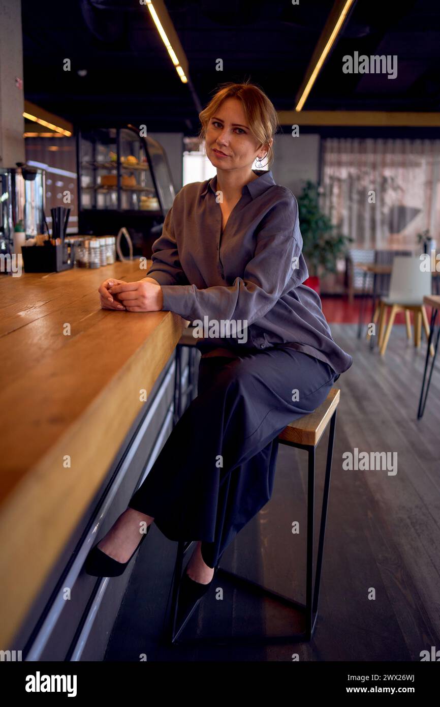 stanca donna di mezza età in un bar dal design neutro, indossa pantaloni a gamba larga e una blusa di seta Foto Stock