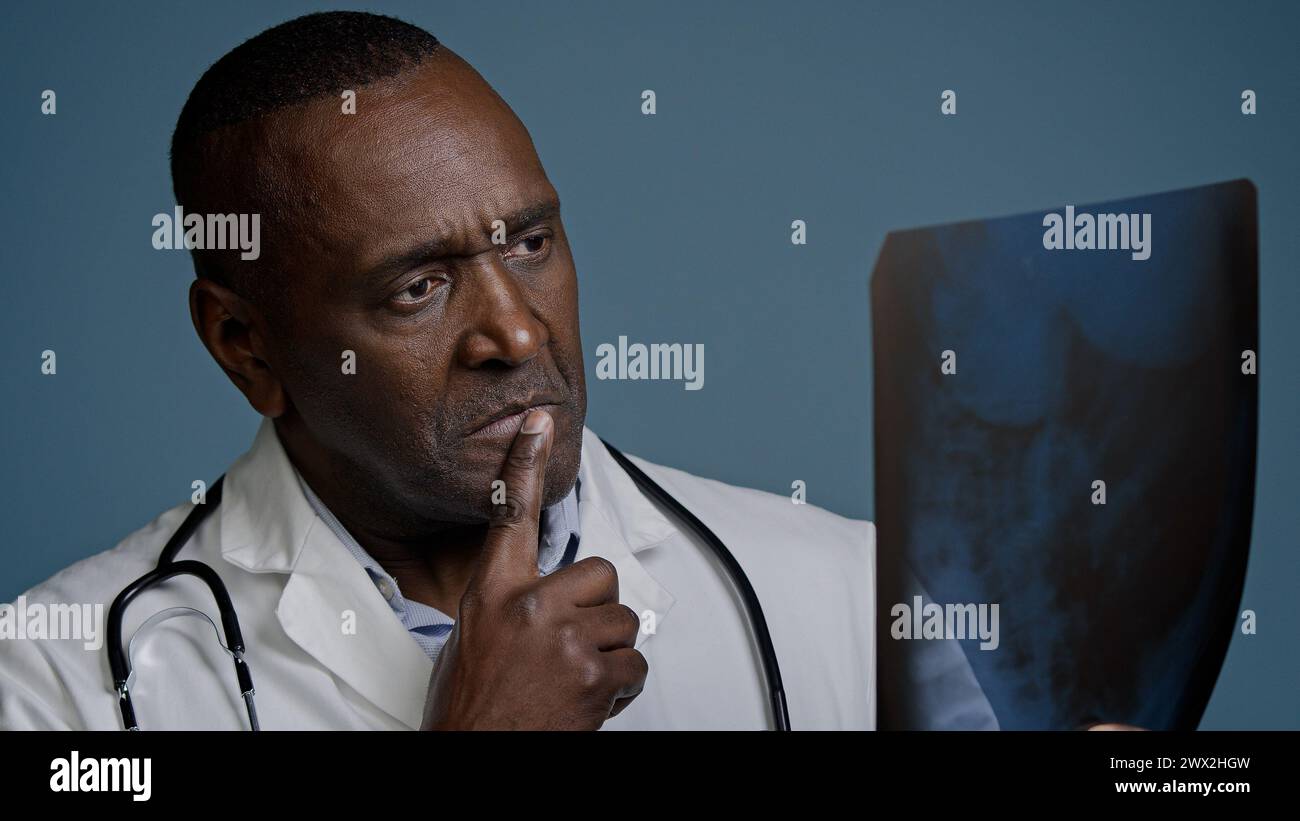 Medico maschio afroamericano pensivo maturo esaminare radiografia in sfondo blu studio uomo chirurgo traumatologo controllare risultati scansione analizzare lesioni su Foto Stock