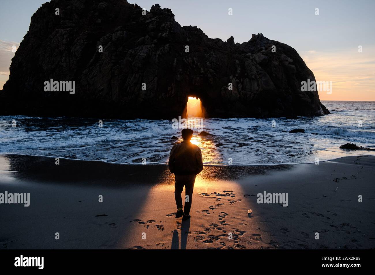 Il tramonto illumina la persona di fronte alla caratteristica Window in the Rock di Keyhole Arch a Pfeiffer Beach a Big Sur sull'Oceano Pacifico in California, Stati Uniti. Foto Stock