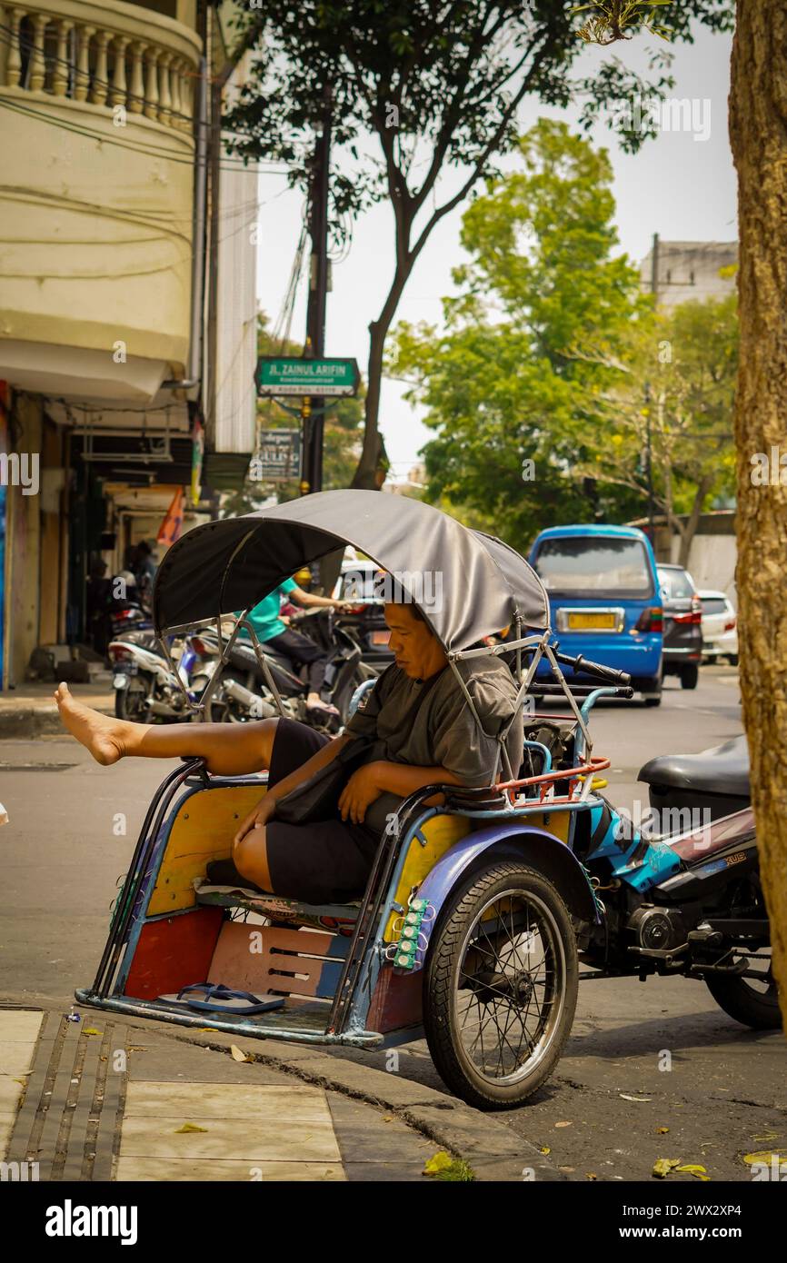 Un uomo è seduto a riposare in un becak (pedicab) parcheggiato sul lato della strada durante il giorno Foto Stock