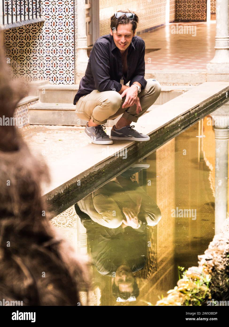 Un uomo di trent'anni si guarda sorridente al riflesso di una fontana mentre accovacciato Foto Stock