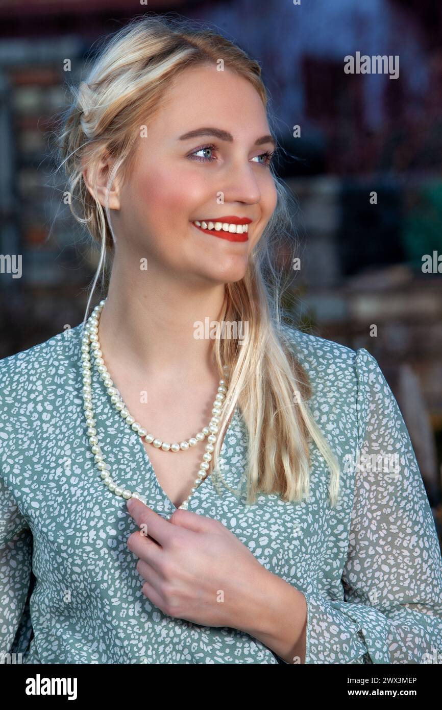 In una calda giornata estiva, una donna bionda con un abito estivo arioso si erge davanti a uno sfondo idilliaco. I suoi capelli cadono dolcemente sulle spalle e. Foto Stock