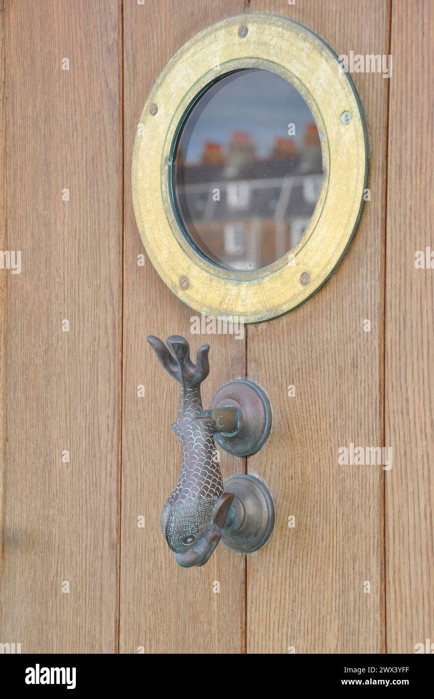 La tradizionale porta in ottone a forma di delfino bussa sotto una finestra a forma di oblò in una porta di legno su questa casa sul porto di Weymouth, Dorset. Foto Stock