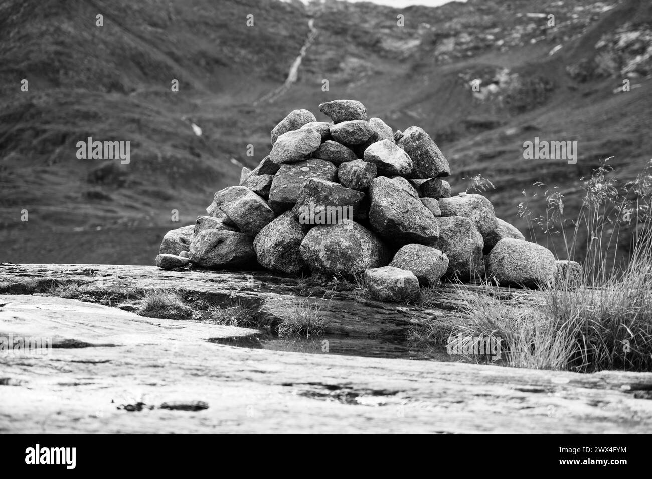 Un cairn accuratamente impilato sorge su un terreno roccioso con vegetazione lussureggiante e sfondo di una cascata. Fotografia in bianco e nero. Foto Stock
