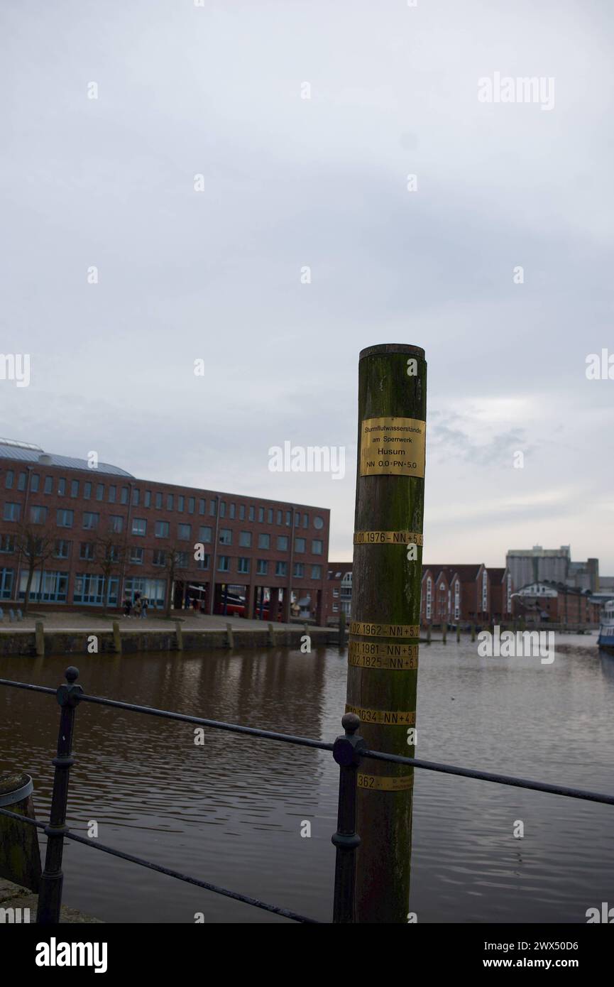 Husum, Schleswig-Holstein Sturmflutpfahl am Ende des Hafenbeckens, der Hafenspitze des Husumer Binnenhafens. Der Pfahl zeigt die Wasserstände nach den wuchtigsten Fluten an, die den Ort maßgeblich prägten und prägen. AM oberen Ende sieht man die Wasserhöhe NN Null am Sperrwerk. CA. 60 cm più scuro die Marke der Sturmflut von 1976. Darunter die von 1962 mit einer Höhe von 5,61 Meter über Normalnull. Kurz darunter die Sturmflut vom 24.11.1981 und die vom 03.02.1825 und 11.10.1634. Aufnahme vom 27.03.2024, Husum, Kreis Nordfriesland, Schleswig-Holstein *** Husum, Schleswig Holstein Storm surge PIL Foto Stock