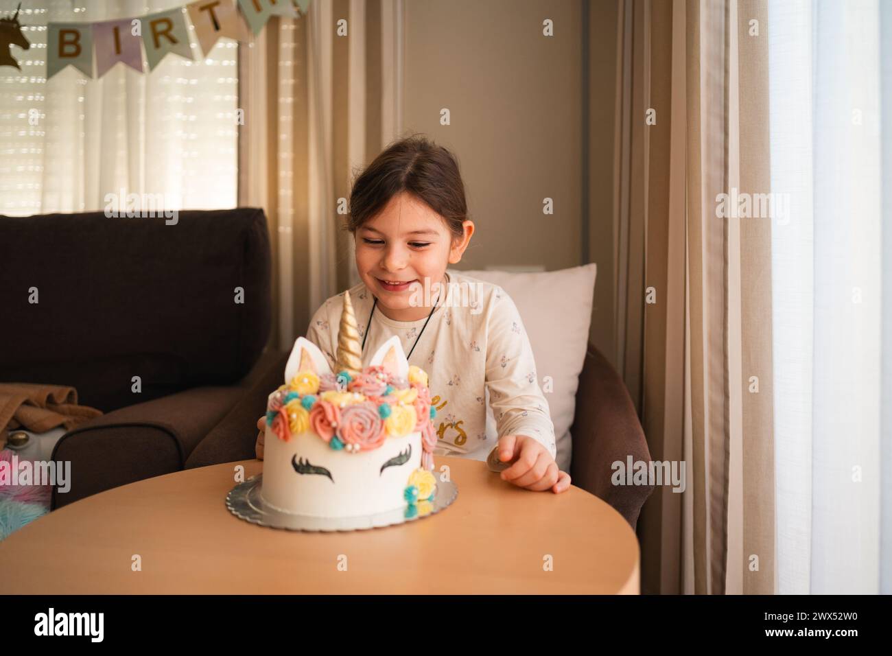 Una gioiosa ragazza di compleanno sorride mentre si allontana per toccare il corno della sua torta da unicorno da sogno, un momento indimenticabile nella sua festa Foto Stock