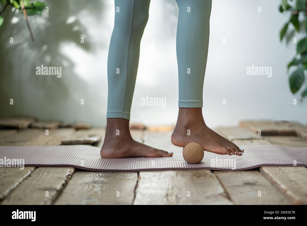 La donna nera sta in piedi sul tappetino da palestra usando la palla massaggiante per tirare fuori i piedi di tensione e dedicare del tempo all'autoassistenza. Foto Stock