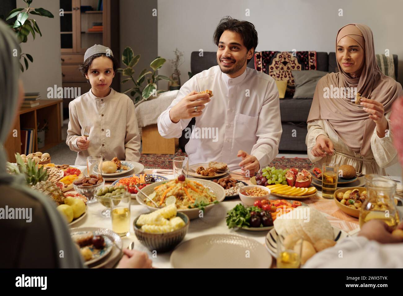 Moderna famiglia musulmana che indossa abiti modesti seduti al tavolo nel soggiorno e che si gode una cena festiva su Eid al-Fitr Foto Stock