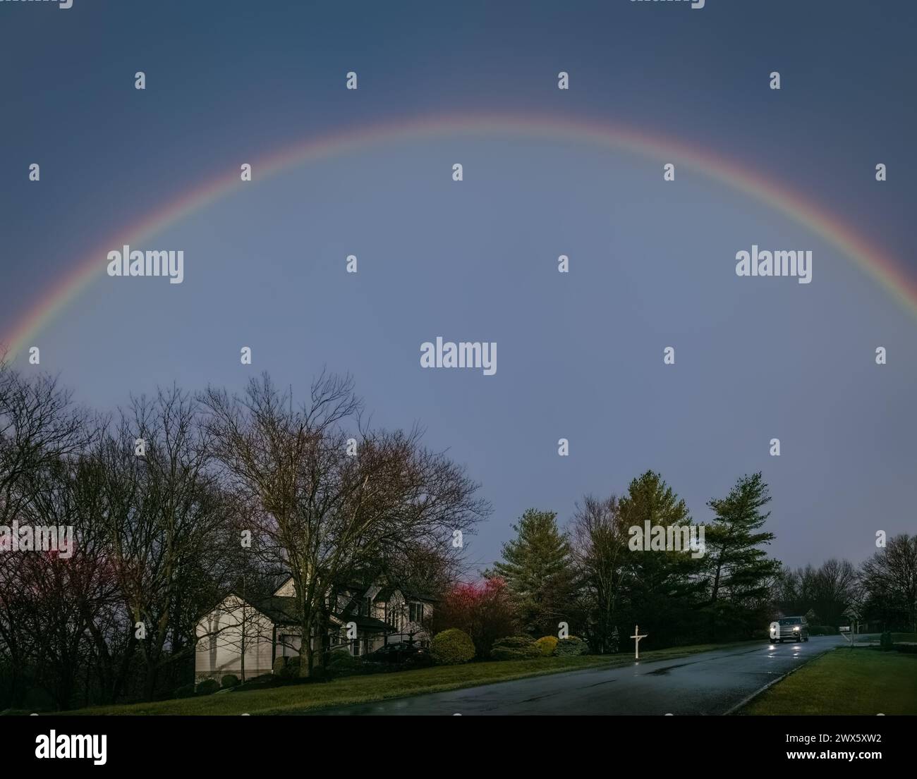Strada suburbana del Midwest la sera delle piogge all'inizio della primavera con l'arcobaleno nel cielo; auto in lontananza Foto Stock