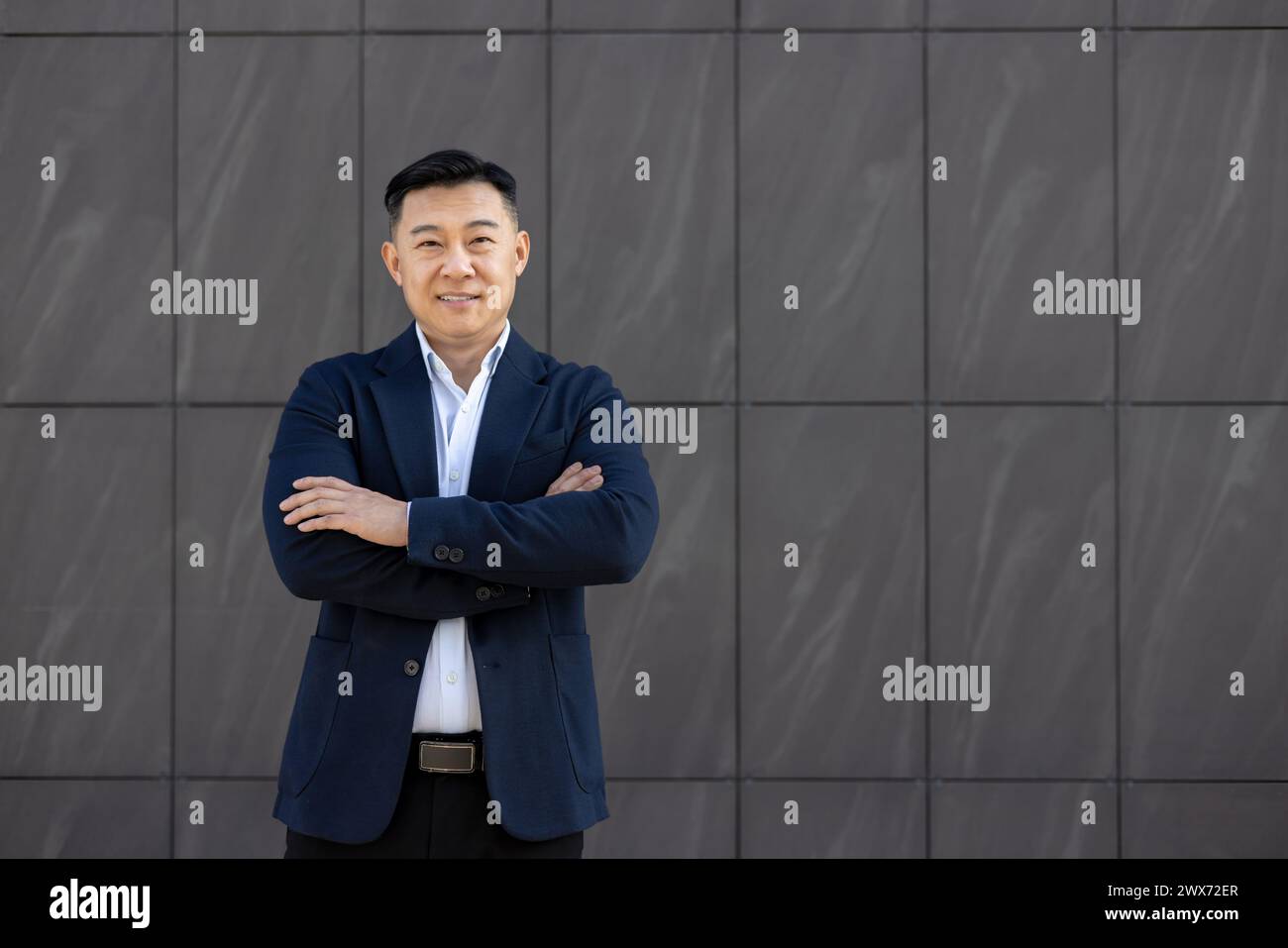 Ritratto di un giovane uomo d'affari asiatico di successo, avvocato in causa in piedi all'esterno sullo sfondo del muro, braccia incrociate sul petto e sorridente con fiducia alla macchina fotografica. Foto Stock