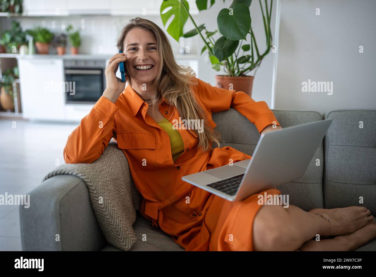 Una donna felice e rilassata sorridente che parla sullo smartphone seduto con il laptop sul divano, dialoga piacevolmente Foto Stock