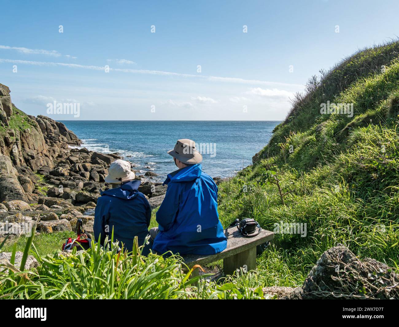 Due camminatori lungo il percorso costiero seduti sul sedile che si affaccia sulla baia di Porthgwarra sul sentiero costiero della Cornovaglia meridionale a marzo, Cornovaglia, Inghilterra, Regno Unito Foto Stock