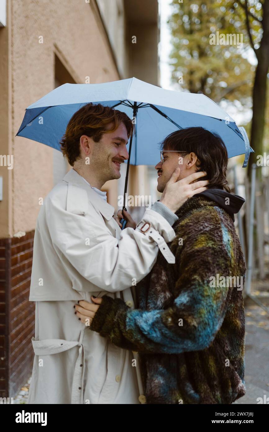 Vista laterale di una giovane coppia gay romantica in piedi sotto l'ombrello sulla strada Foto Stock