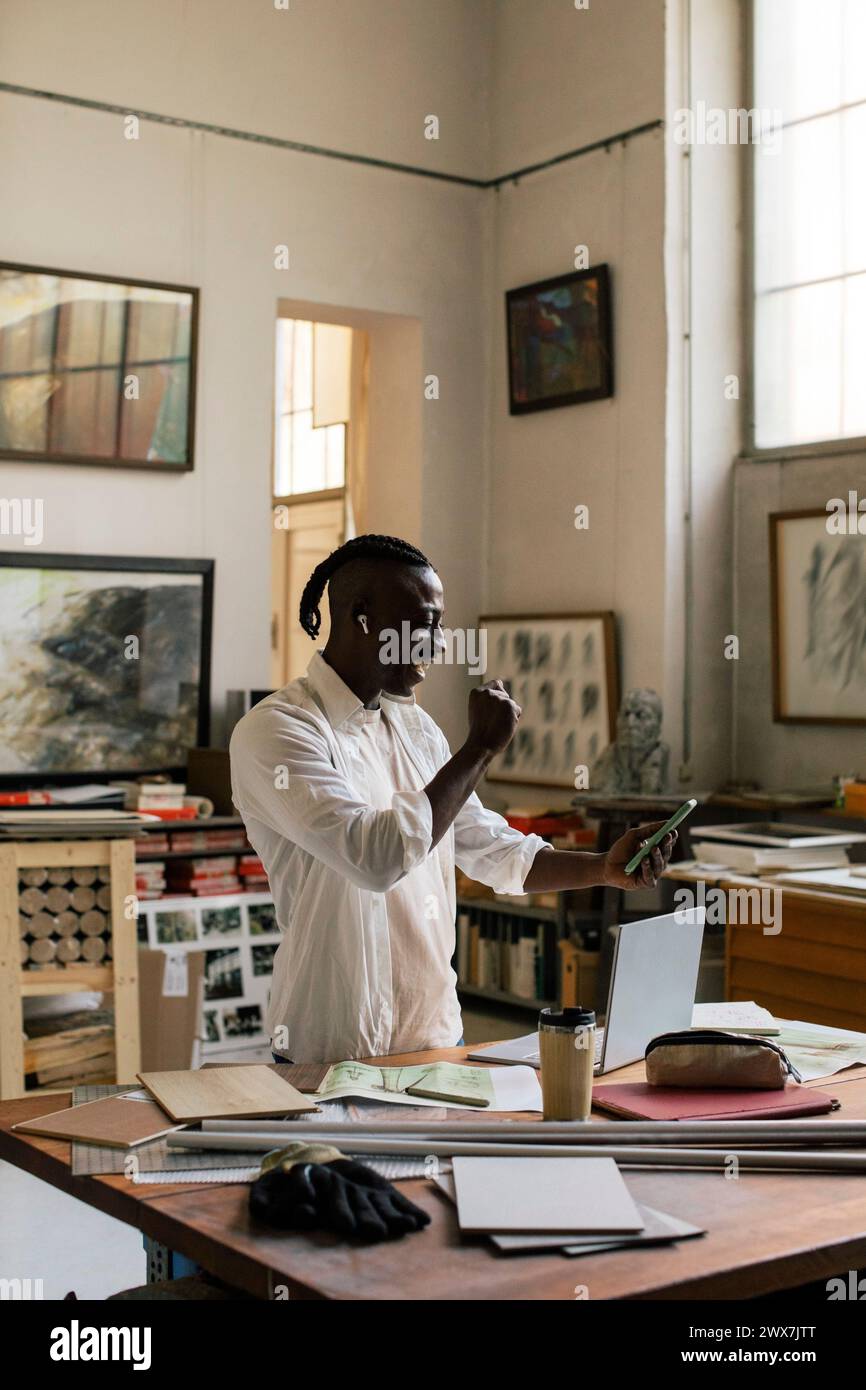 Uomo d'affari sorridente che stringe il pugno e utilizza lo smartphone mentre è in officina Foto Stock