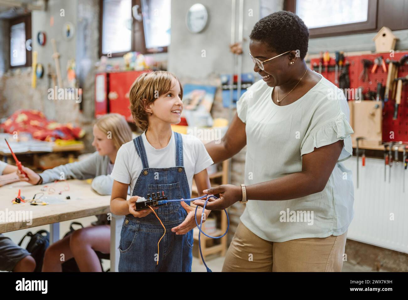 Insegnante sorridente che incoraggia la studentessa a tenere parte elettrica presso un laboratorio tecnologico a scuola Foto Stock