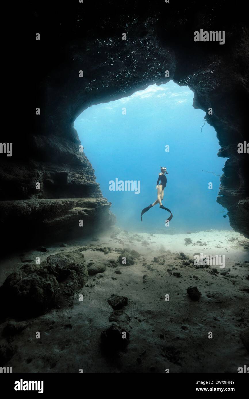 Una giovane femmina che si tuffa liberamente in una porta di una grotta nelle limpide acque blu delle Hawaii incorniciate da pareti ombrose della grotta. Rilascio modelli disponibile Foto Stock