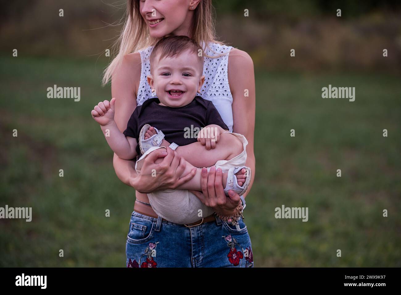 La giovane donna sorride, tiene il bambino tra le braccia, in piedi in un campo lussureggiante con il soffice bagliore della luce serale. Il concetto di tattilità, conn materno Foto Stock