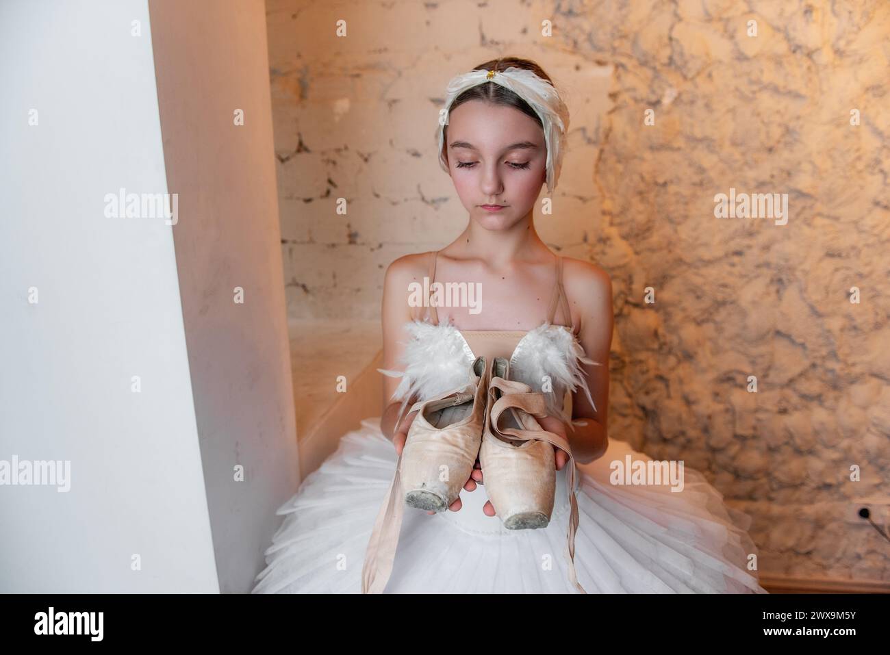 Giovane ballerina in tutù bianco, accessorio tiara con capelli piumati, regge con cura le scarpe pointe indossate, evocando il senso di dedizione e la quie Foto Stock