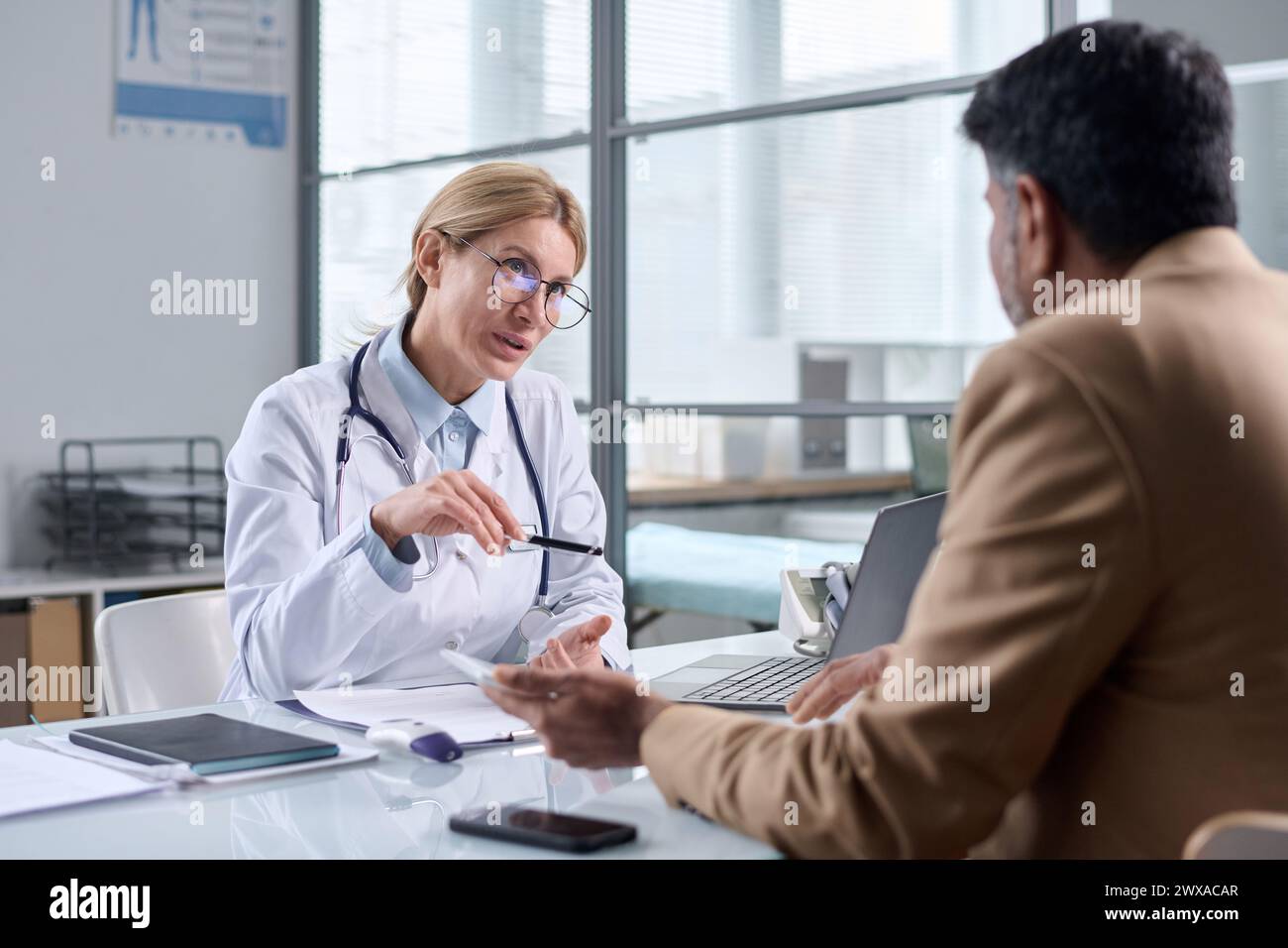 Ritratto di medico donna maturo che indossa occhiali e parla con il paziente attraverso il tavolo in clinica medica Foto Stock