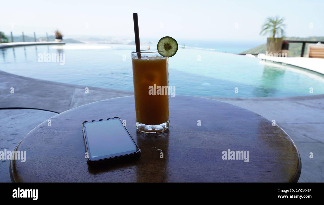 Succo di mango in un alto vetro trasparente con un cuneo di lime per la decorazione e paglia di legno con smartphone e una piscina come sfondo Foto Stock