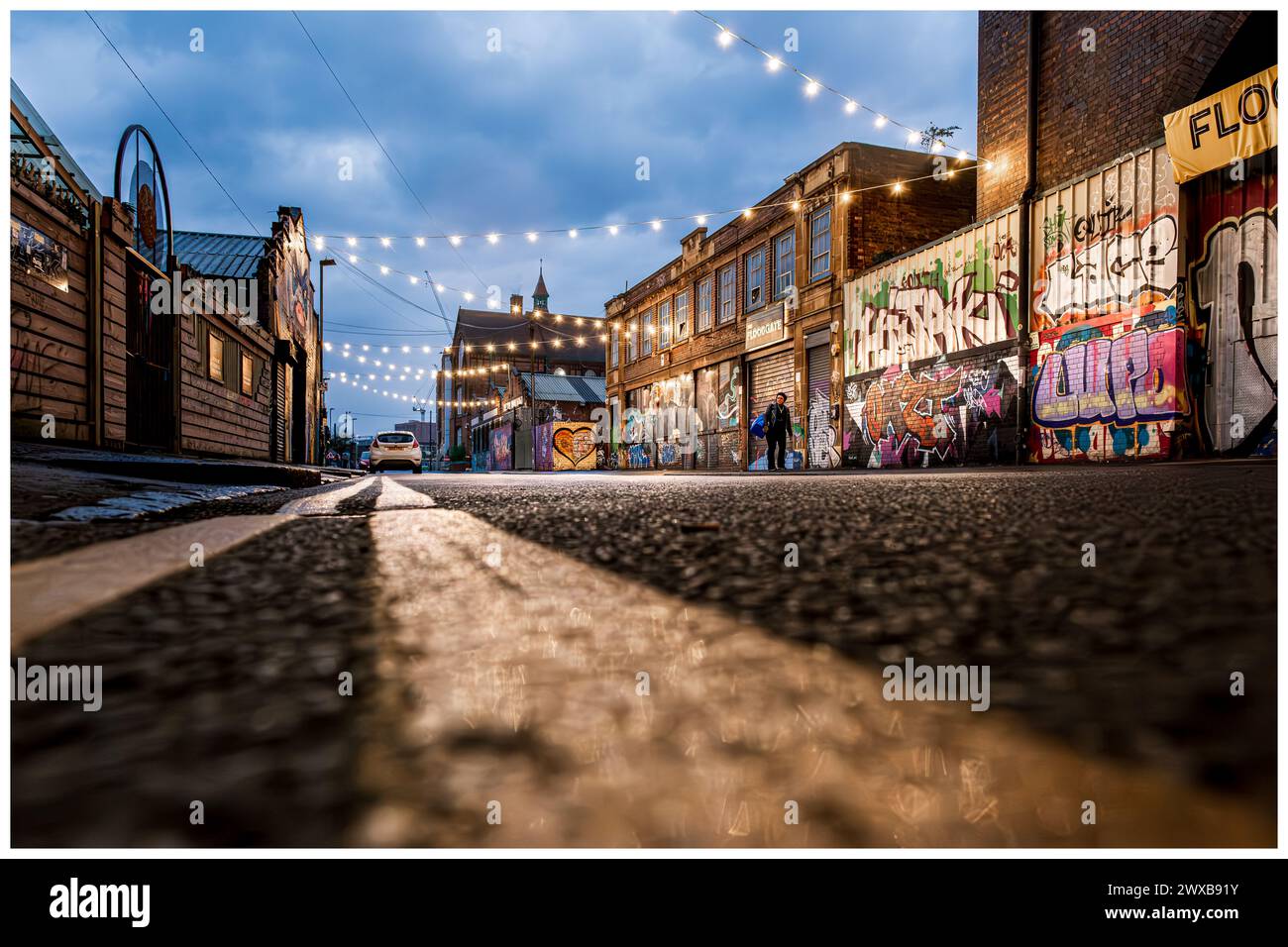 vista a livello della strada lungo la strada secondaria urbana al crepuscolo con graffiti e catene luminose decorative. Una persona che cammina Foto Stock